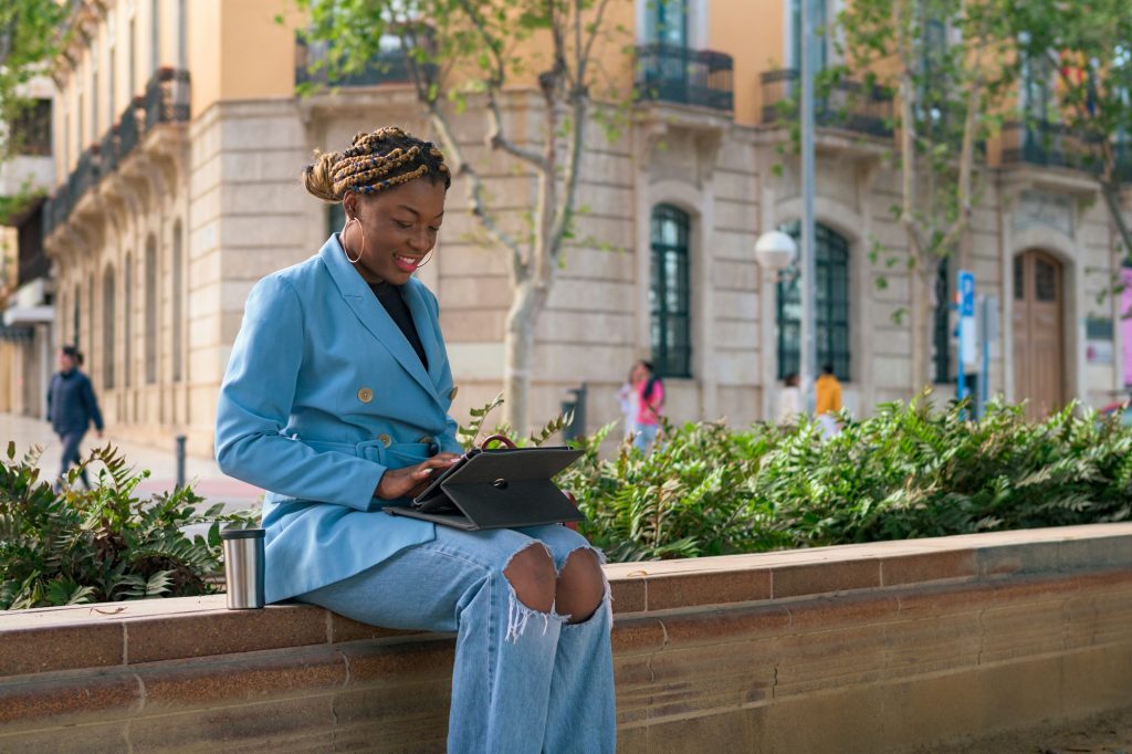 Black business woman is working remotely in a park of a city with a touch laptop